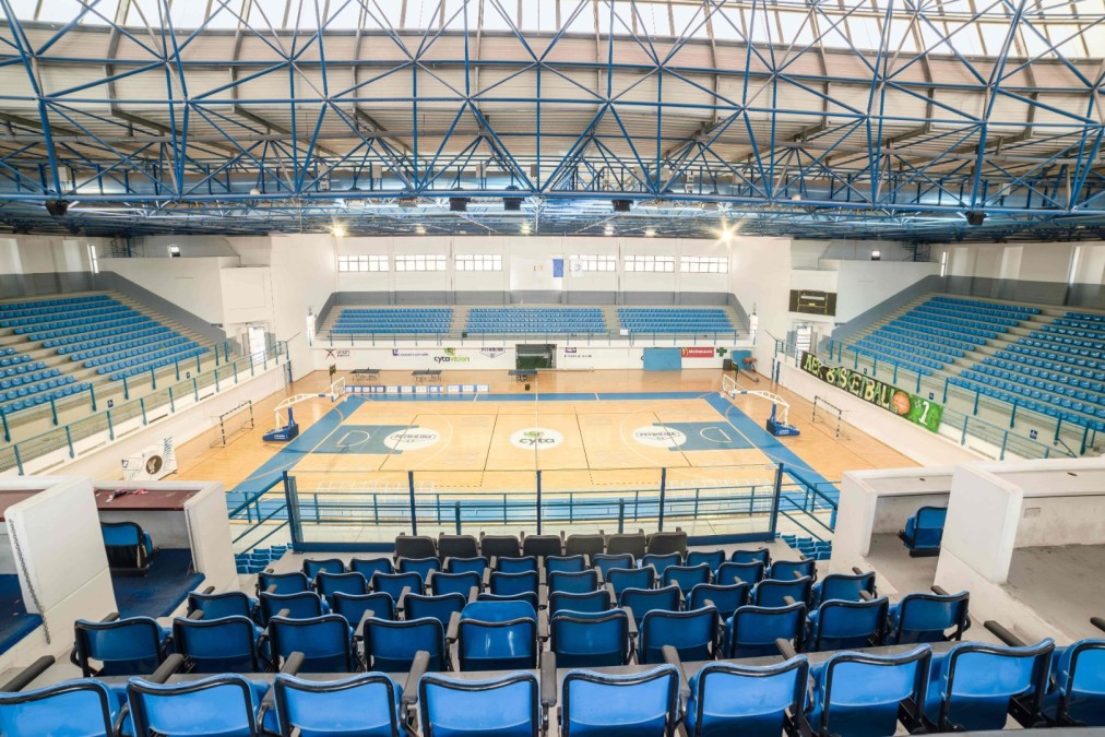 Indoor-Sportarena mit Spielfeldmarkierungen, Körben und blauen Sitzreihen.