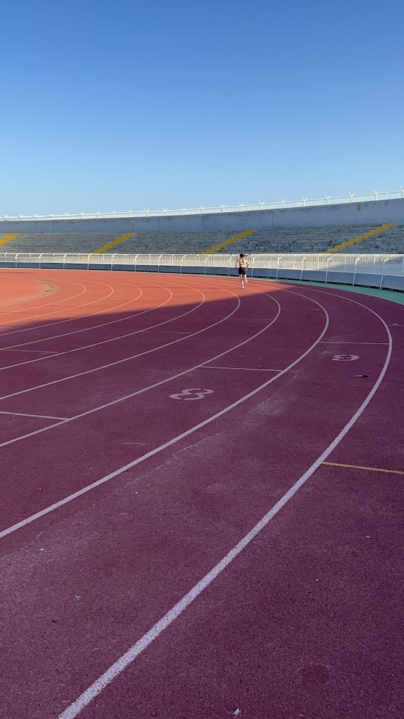 Gebogene Leichtathletikbahnen mit einer laufenden Person und leeren Tribünen im Hintergrund.