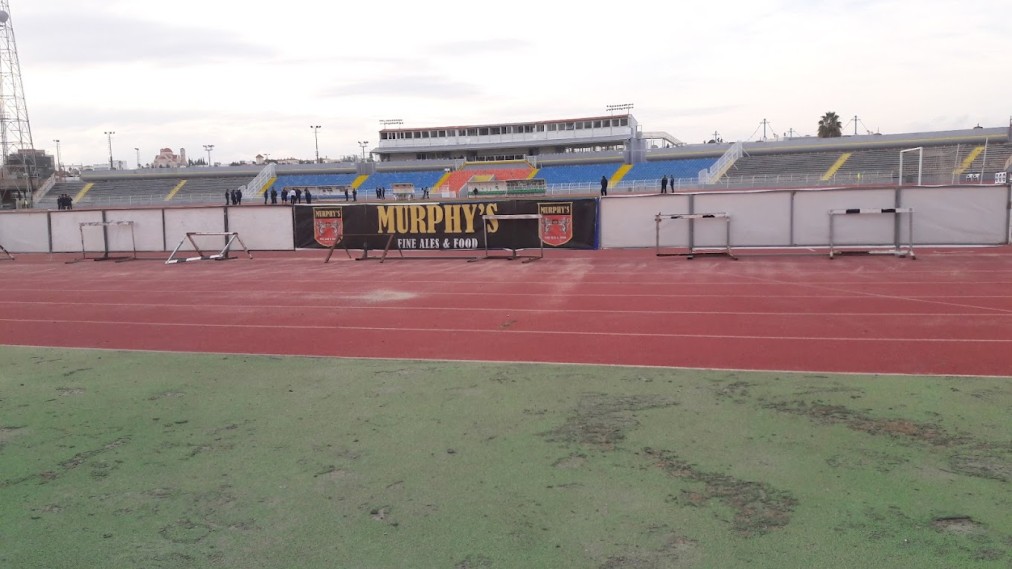Blick von der Laufbahn auf ein Stadion mit Tribünen, Flutlichtmasten und Spielfeldabsperrungen.