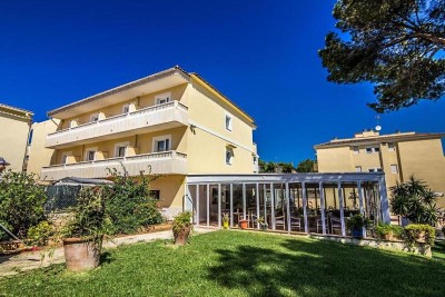 Hotelaußenansicht mit drei Etagen, Garten und überdachtem Glas-Loungebereich.