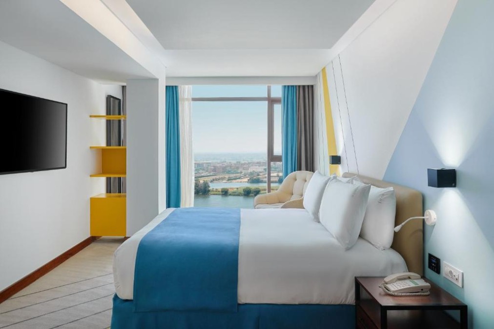 Hotelzimmer mit blauer Tagesdecke, Fernseher an der Wand und Sessel, großes Fenster mit Blick auf Fluss und Stadt