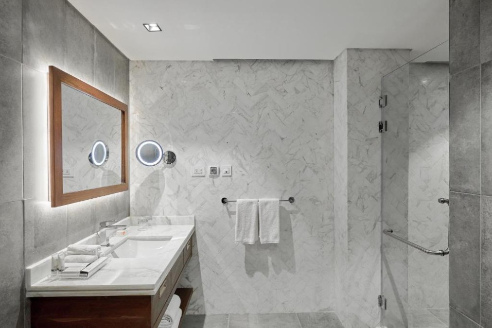 Hotelbadezimmer mit grauen Fliesen, Marmorwänden, Waschtisch, beleuchtetem Spiegel, Handtuchhalter und Glasdusche