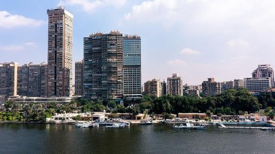 Flussufer-Skyline mit Hochhäusern, Bäumen und anlegenden Booten unter einem teils bewölkten blauen Himmel