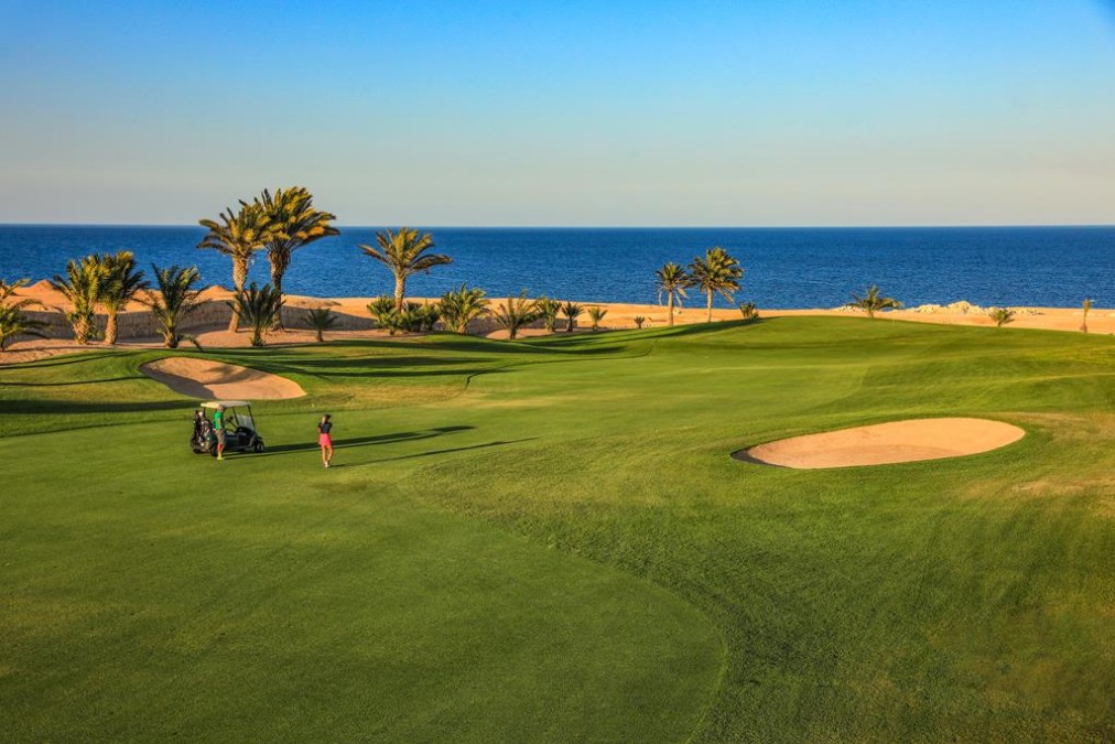 Golfeur près d'une voiturette sur un fairway en bord de mer avec palmiers et bunkers de sable.