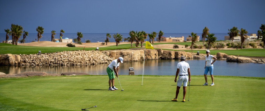 Trois golfeurs sur un green de putting près d'un obstacle d'eau, avec palmiers et mer en arrière-plan.