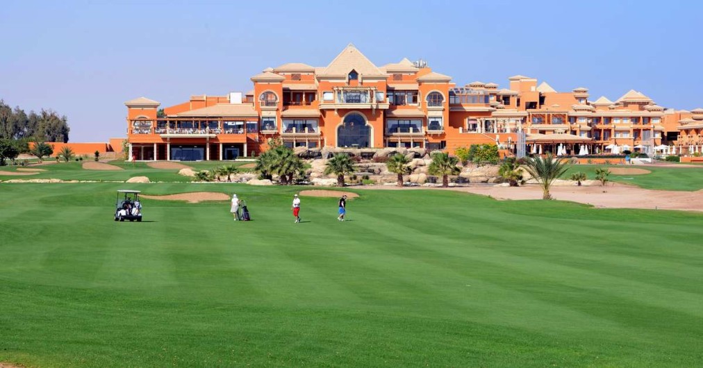 Golfeurs et voiturette sur un fairway devant un grand bâtiment de club-house orange.