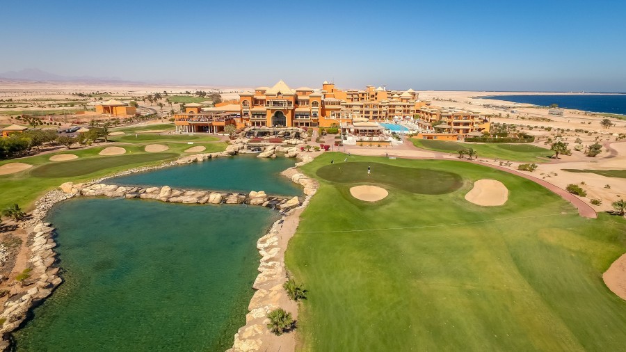 Vue aérienne d'un complexe à côté d'un trou de golf avec bunkers et lagon turquoise, mer au-delà.