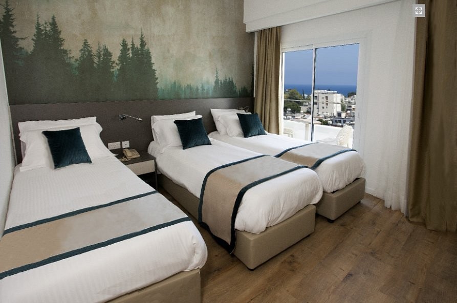 Chambre triple avec trois lits simples, un mur orné d'une fresque forestière et des portes-fenêtres avec vue sur la ville et la mer.