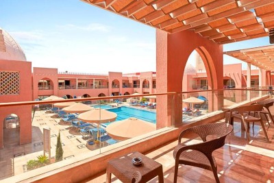 Vue depuis le balcon sur la piscine du complexe avec parasols et architecture rouge en arcades