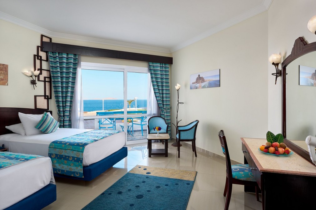 Chambre d'hôtel lumineuse avec lits jumeaux, décor bleu et balcon offrant vue sur la mer, idéale pour l'hébergement d'un camp sportif