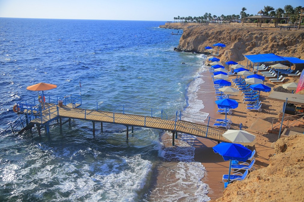 Plage de sable avec parasols bleus, transats et jetée en bois s'étendant dans la mer claire