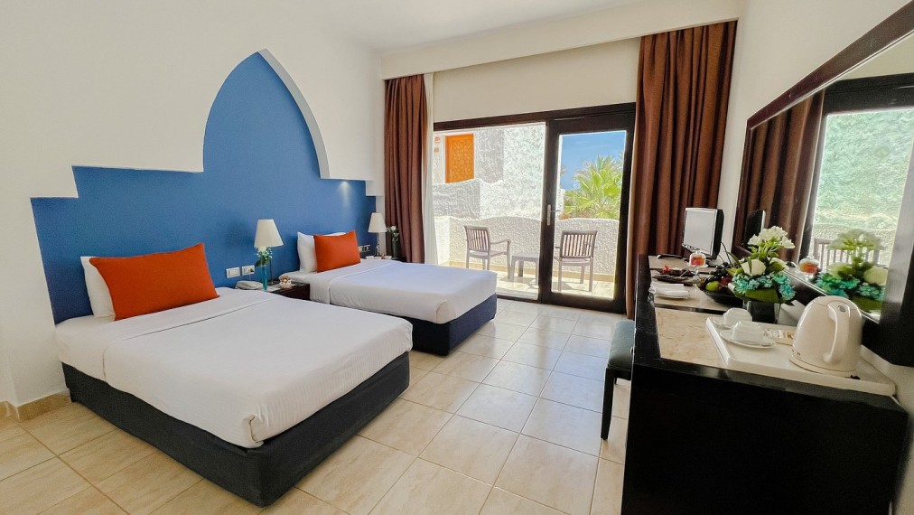 Chambre lumineuse à deux lits avec touches bleues et orange, balcon privé et coin salon extérieur avec vue sur la station