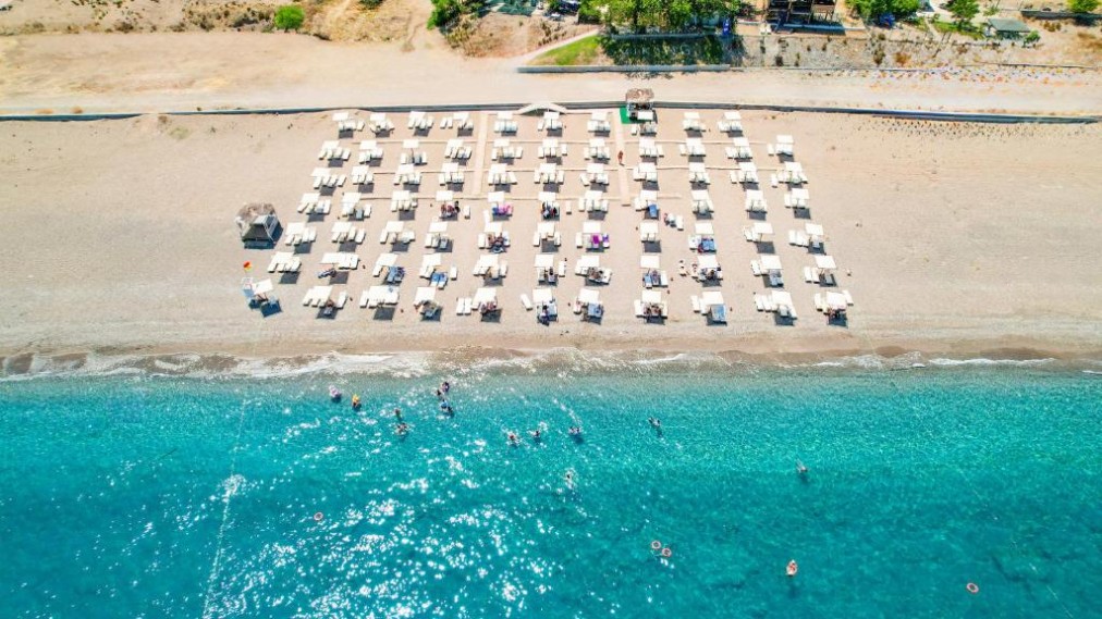 Vue aérienne de rangées de transats sur la plage au bord d'une mer turquoise ; baigneurs près du rivage.