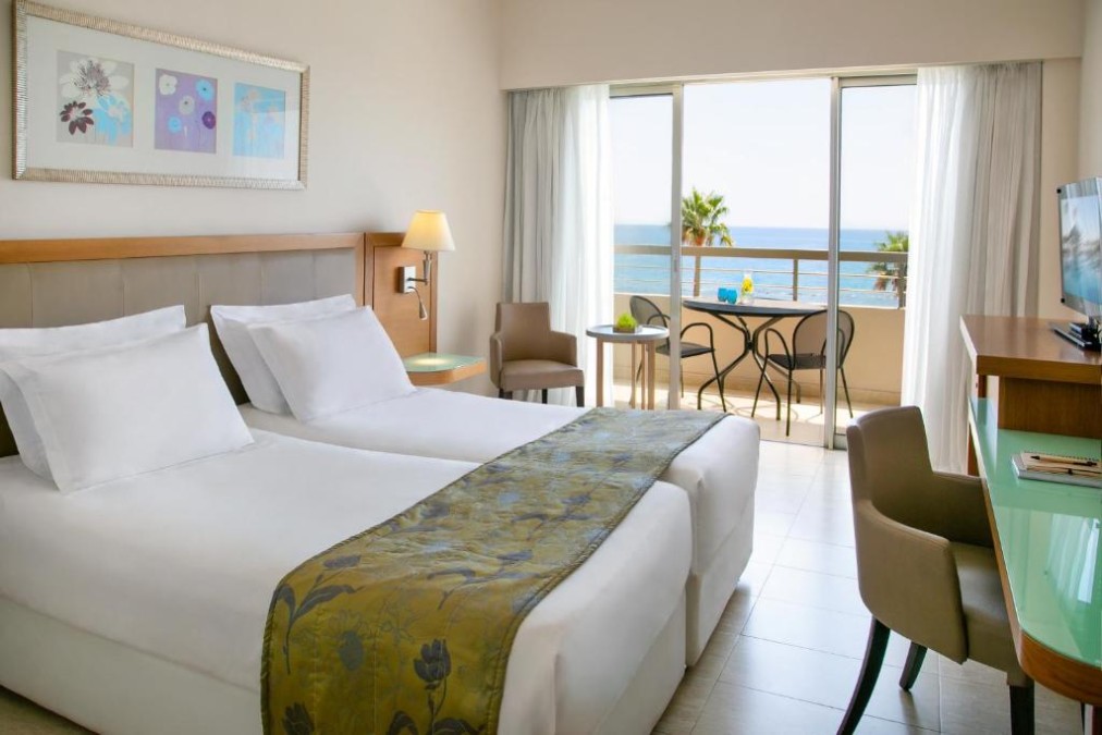 Chambre d'hôtes avec balcon vue mer, lits jumeaux, bureau et fauteuil.